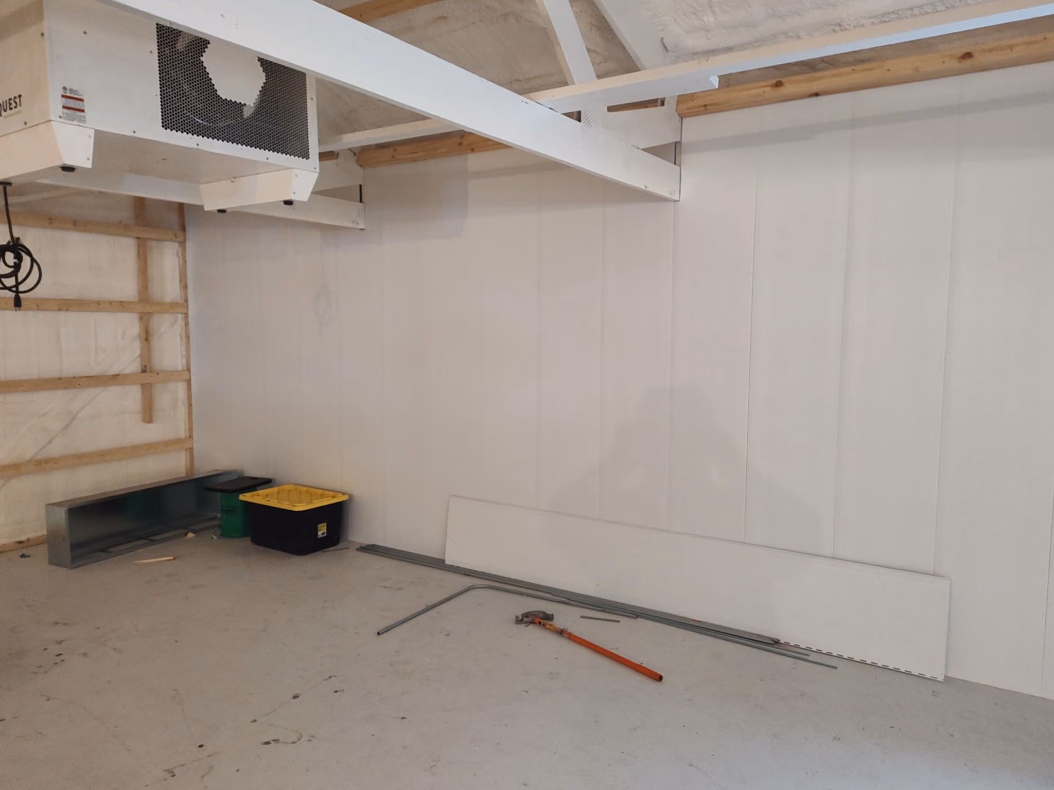 Empty room with white walls, exposed beams, a pipe wrench, metal bars, and plastic containers on the floor.