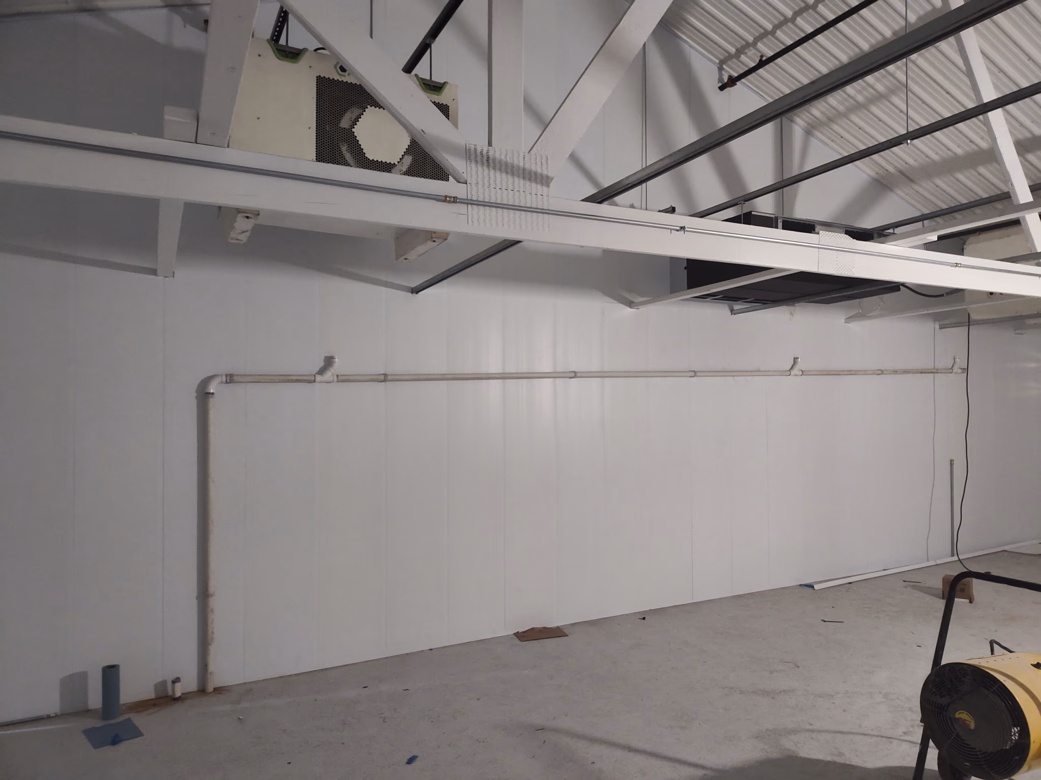 Interior of an empty industrial space with white paneled wall, exposed ceiling beams, and pipes.