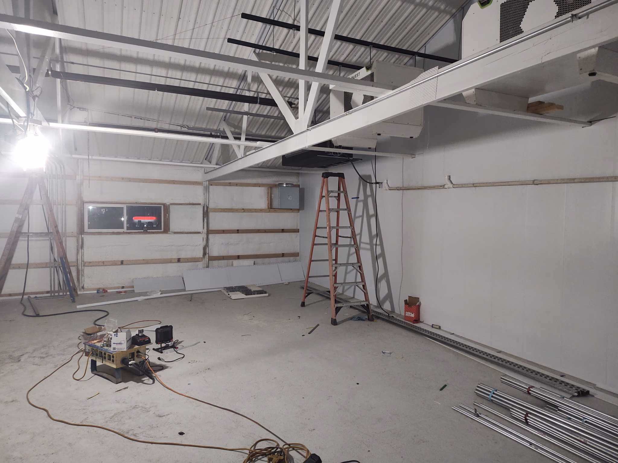 Empty room under construction with ladders, metal pipes, cables, and insulation on walls and ceiling.
