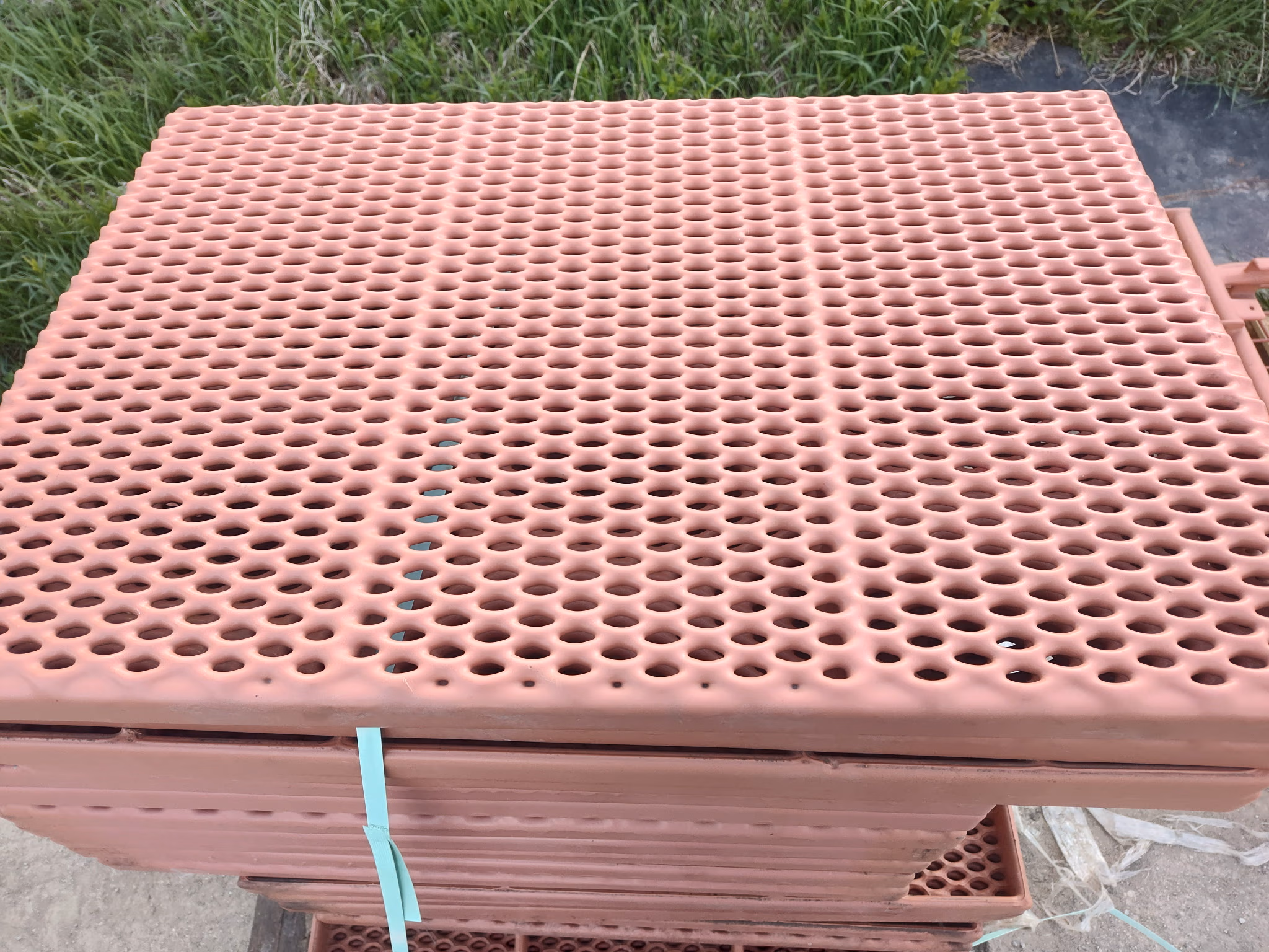 Stack of pink perforated plastic sheets tied with a green strap outdoors on concrete and grass.